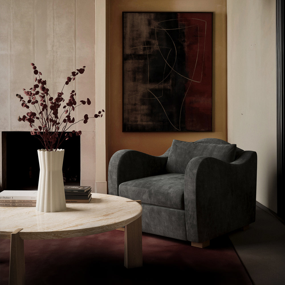 Lemieux et Cie Elodie Lounge Chair in charcoal upholstery with soft arms, styled in a sitting room with stone table, warm ochre walls, and abstract art.