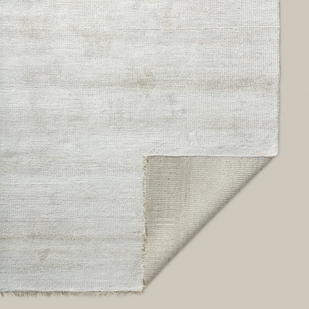 Lemieux et Cie Rochefort Handmade Wool Rug in ivory, woven rug shown with corner fold revealing textured surface and fringe edge on neutral backdrop.