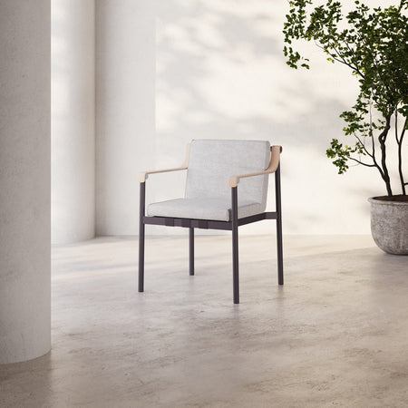 Lemieux et Cie Hudson Dining Arm Chair with light upholstery and dark slender frame, shown on concrete flooring beside white column and potted tree in soft daylight.