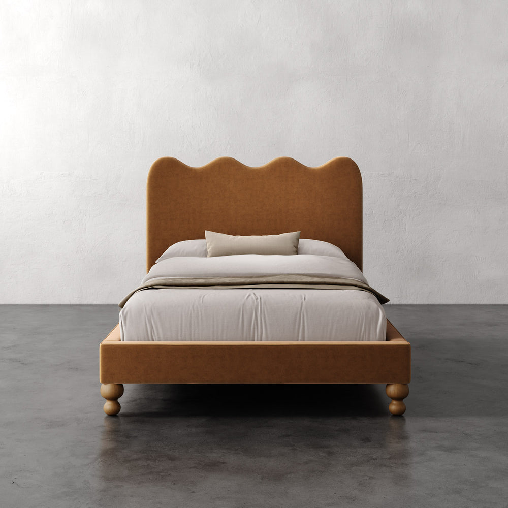 Lemieux et Cie Celine Bed with scalloped headboard and turned legs in warm caramel upholstery, styled with neutral linens on concrete floor against white plaster wall.