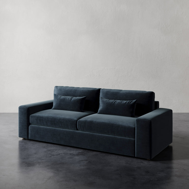 Lemieux et Cie Lacroix Sofa in deep slate blue velvet, low-profile sofa with bolster pillows, shown on dark concrete flooring against textured plaster wall.