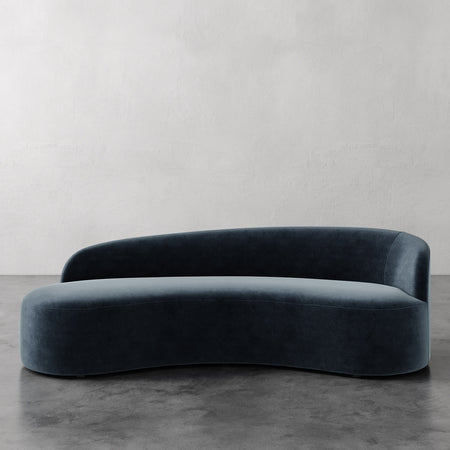Lemieux et Cie Colette Right Arm Sofa in deep teal velvet, sculptural curved sofa with asymmetric silhouette, shown on concrete floors against textured plaster walls.
