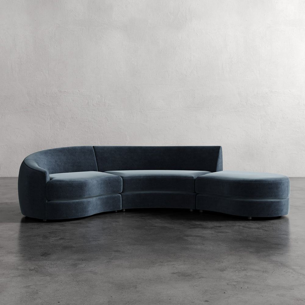Lemieux et Cie Rulmann 3-Piece Left Sectional, curved sofa in deep teal upholstery, shown on concrete floor against textured plaster walls.