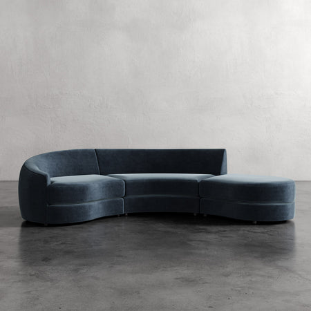 Lemieux et Cie Rulmann 3-Piece Left Sectional, curved sofa in deep teal upholstery, shown on concrete floor against textured plaster walls.