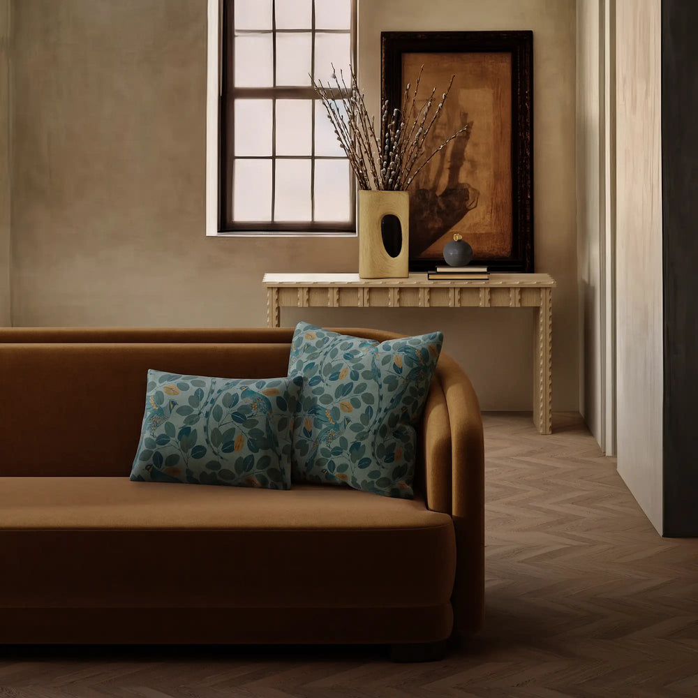 Lemieux et Cie Modern Peacock Velvet Pillow in teal and copper, decorative throw pillow on a warm sofa in a room with herringbone wood floors and plaster walls.