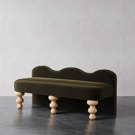 Lemieux et Cie Celine Bench in olive green, sculptural low-profile bench with undulating backrest and turned legs, shown on dark stone floor against white plaster wall.