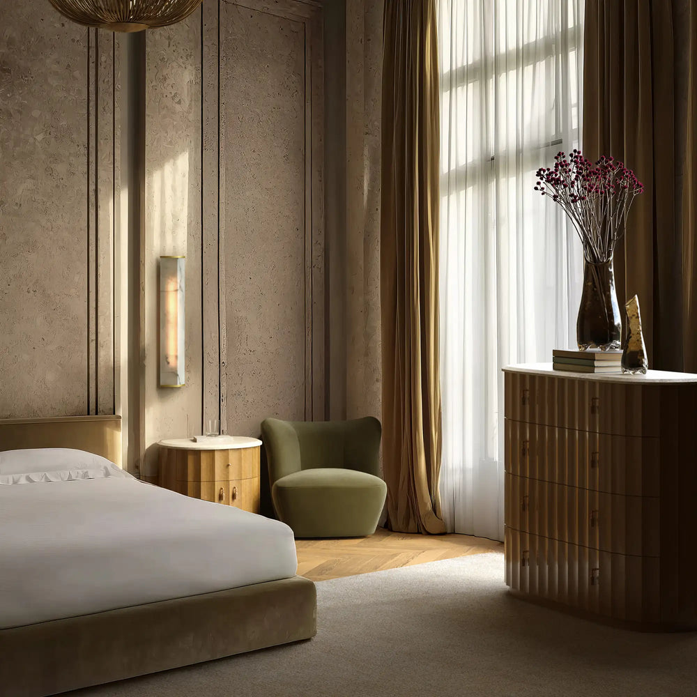Lemieux et Cie Ondara Fluted 5-Drawer Dresser in champagne eucalyptus with honed travertine top, styled in a bedroom with textured stone walls, warm drapery and natural light.