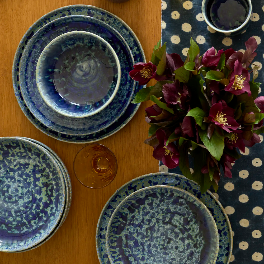 Lemieux et Cie Hanami Stoneware Dinnerware Set in indigo with reactive glaze, nested plates and bowls on wood table with fresh flowers and patterned runner.
