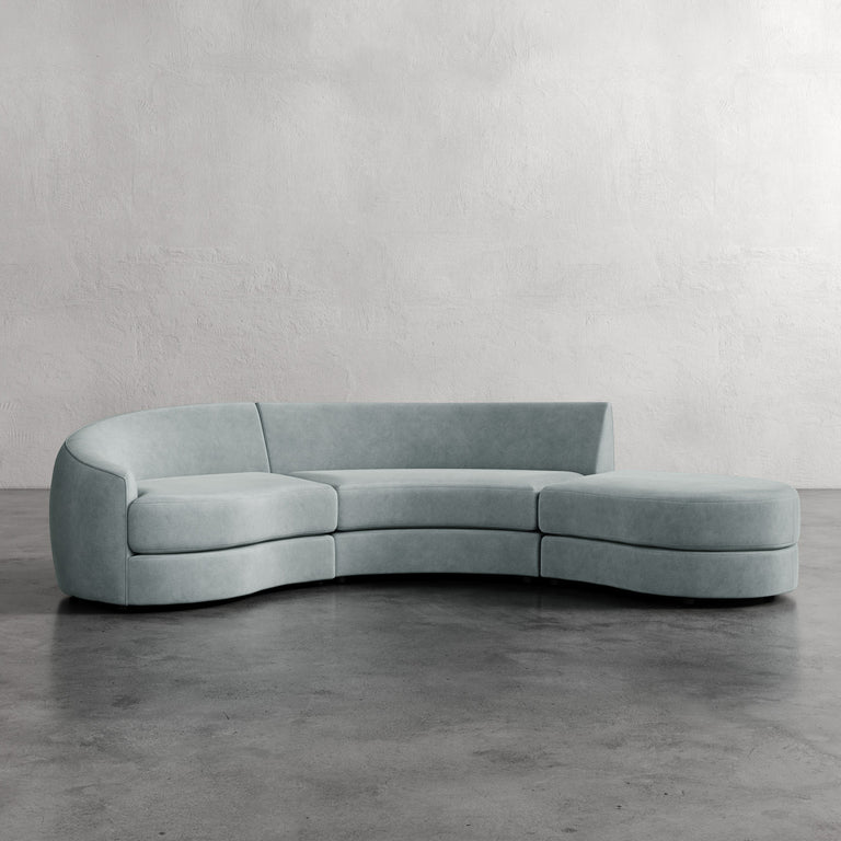 Lemieux et Cie Rulmann 3-Piece Left Sectional in soft blue upholstery, curved modular sofa on concrete floor against textured plaster walls.