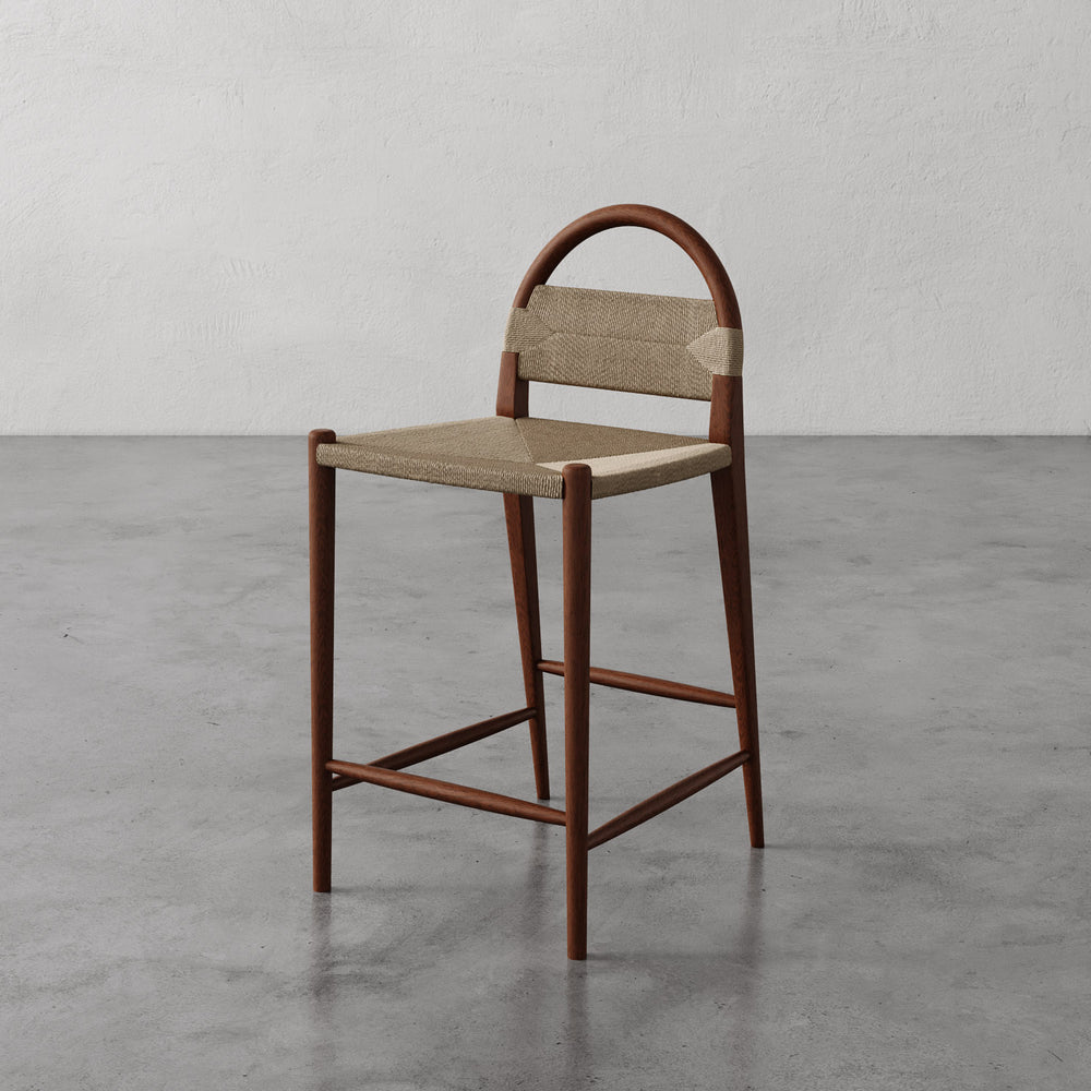 Lemieux et Cie Pernelle Counter Stool with arched backrest and woven seat, warm-toned frame on concrete floor against textured plaster walls.