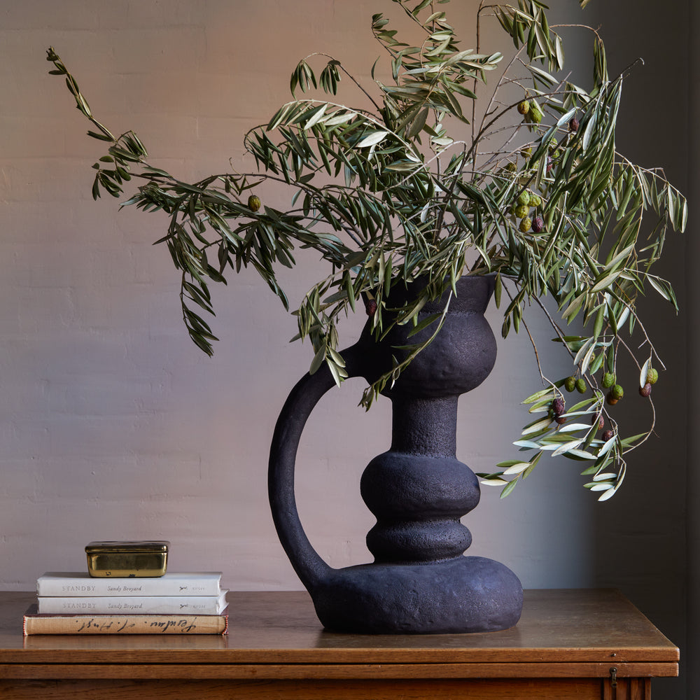 Lemieux et Cie Remi Vase in black handcrafted ceramic, sculptural organic vessel with stacked spherical form holding olive branches, styled on wood table with neutral plaster walls.