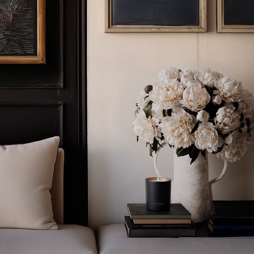 Lemieux et Cie Figue Marseillaise candle in matte black vessel, styled on stacked books beside ivory vase with white peonies in a sitting room with dark paneled walls.