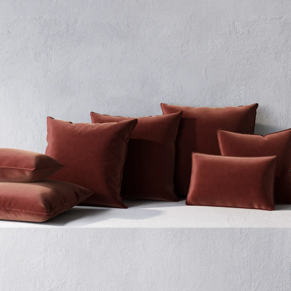 Lemieux et Cie Throw Pillows in terracotta with welted edges, soft pillows in multiple sizes arranged against textured white plaster wall.