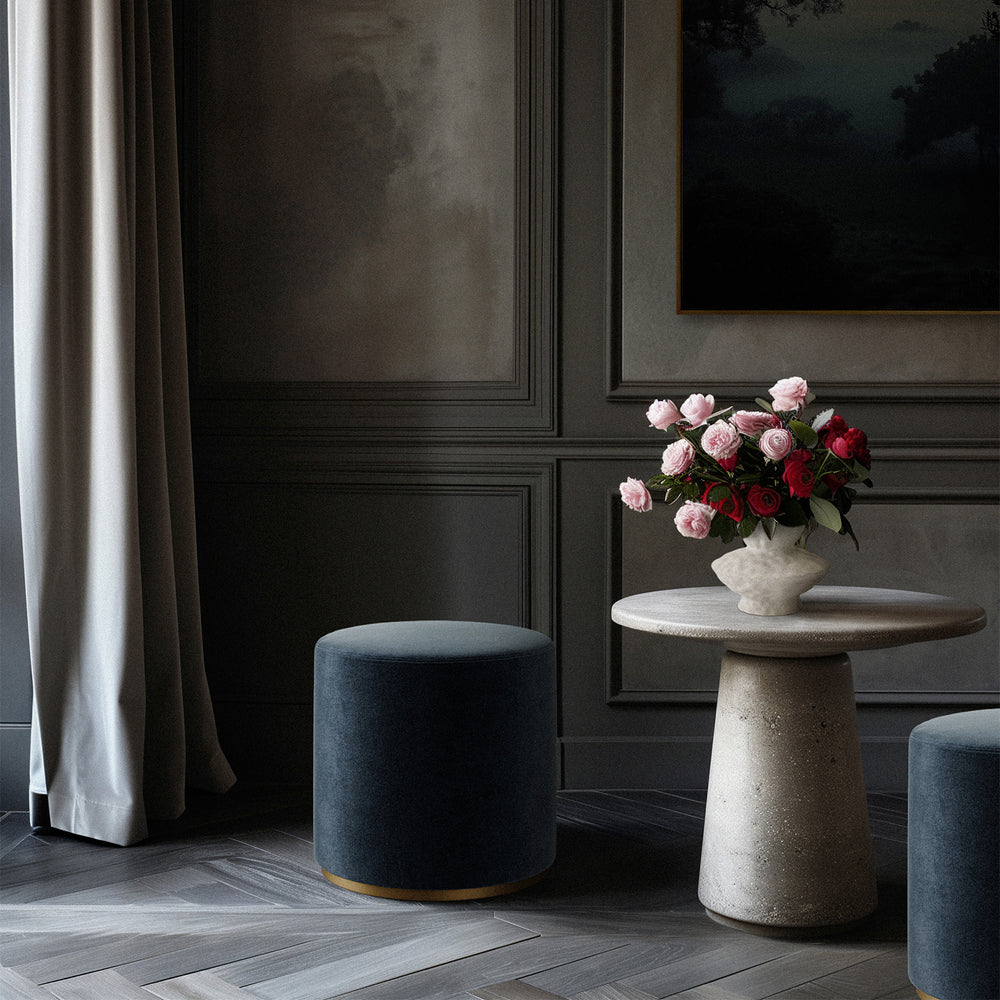 Lemieux et Cie Sophie Round Ottoman in charcoal upholstery with dark cylindrical ottomans and stone side table, styled in paneled sitting room with parquet floors.
