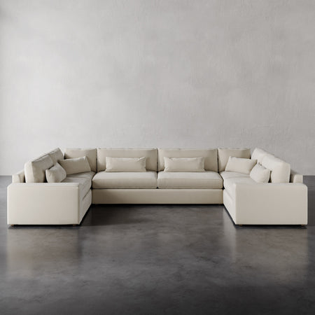 Lemieux et Cie Lacroix U-Sectional in neutral upholstery, modular sofa with deep cushions and bolster pillows, shown on polished concrete floor against white plaster walls.