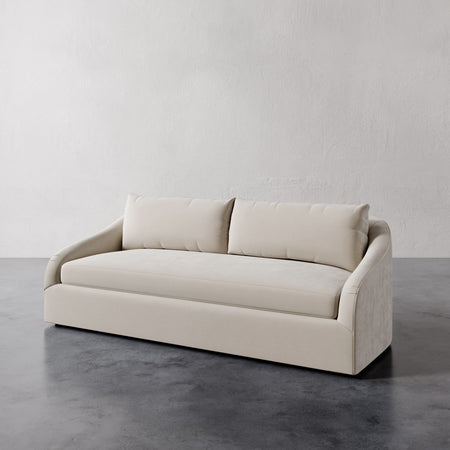 Lemieux et Cie Rousseau Sofa in ivory upholstery, curved-arm sofa with single bench cushion, on dark concrete floor against neutral plaster wall.