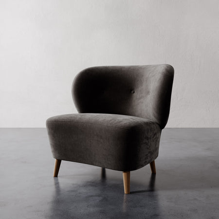 Lemieux et Cie Tourenne Chair in charcoal upholstery, rounded sculptural slipper chair on tapered wood legs, shown on polished concrete floor against white plaster wall.