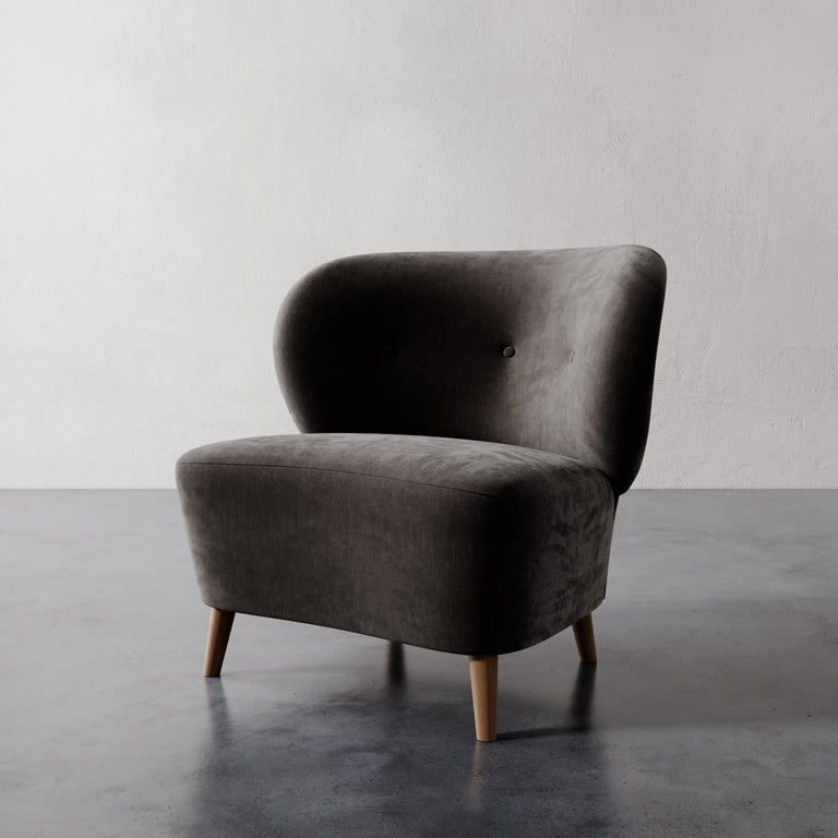 Lemieux et Cie Tourenne Chair in charcoal upholstery, rounded sculptural slipper chair on tapered wood legs, shown on polished concrete floor against white plaster wall.