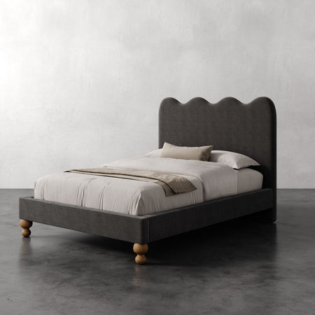 Lemieux et Cie Celine Bed with scalloped headboard in charcoal upholstery and turned legs, styled with neutral linens on dark concrete floor.