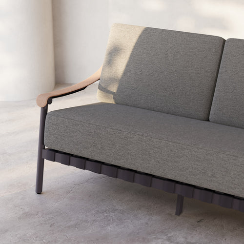 Lemieux Et Cie Hudson 3-Seat Sofa An outdoor sofa with gray fabric cushions, faux all-weather leather arms, and a powder coated aluminum frame, set in a contemporary indoor-outdoor setting with a tree in the background.