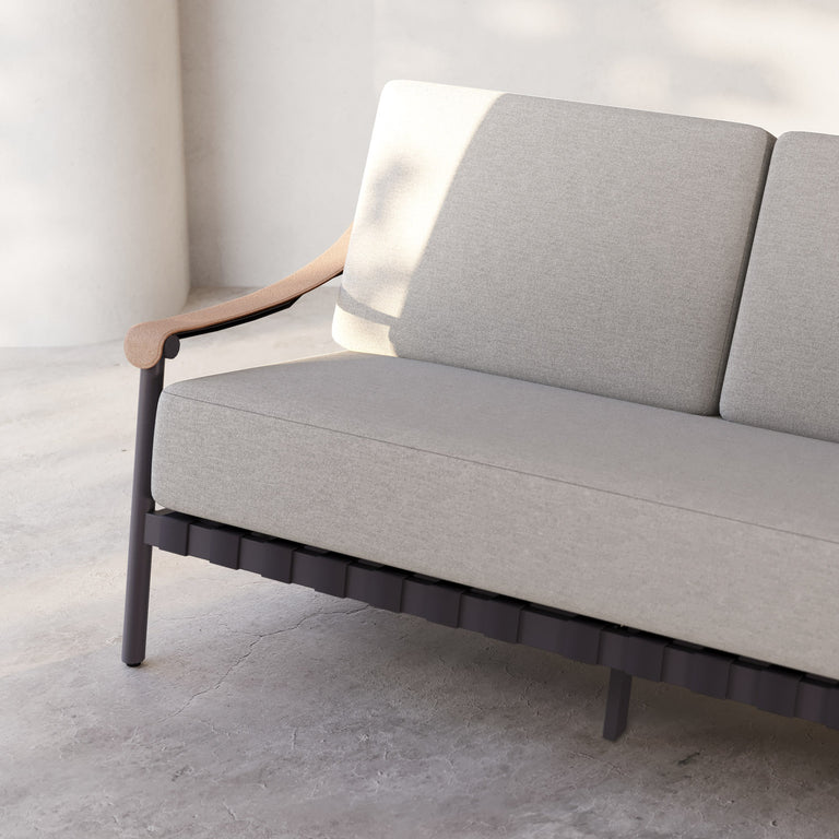 Lemieux Et Cie Hudson 3-Seat Sofa An outdoor sofa with gray fabric cushions, faux all-weather leather arms, and a powder coated aluminum frame, set in a contemporary indoor-outdoor setting with a tree in the background.