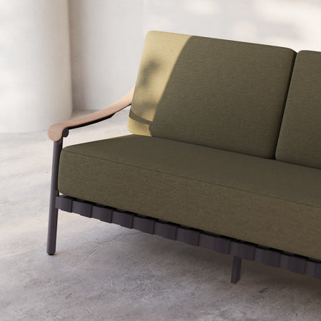 Lemieux et Cie Hudson Lounge Chair in olive upholstery with curved arms and slatted base, shown on concrete floor against white walls in soft daylight.