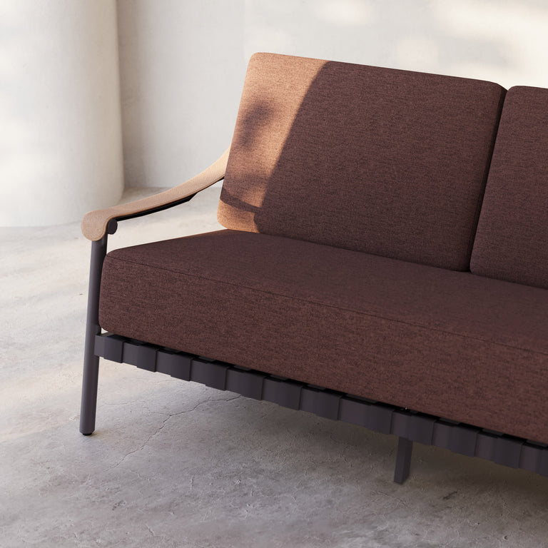 Lemieux Et Cie Hudson Dining Chair swatches_0008-mulberry_copy An outdoor sofa with gray fabric cushions, faux all-weather leather arms, and a powder coated aluminum frame, set in a contemporary indoor-outdoor setting with a tree in the background.