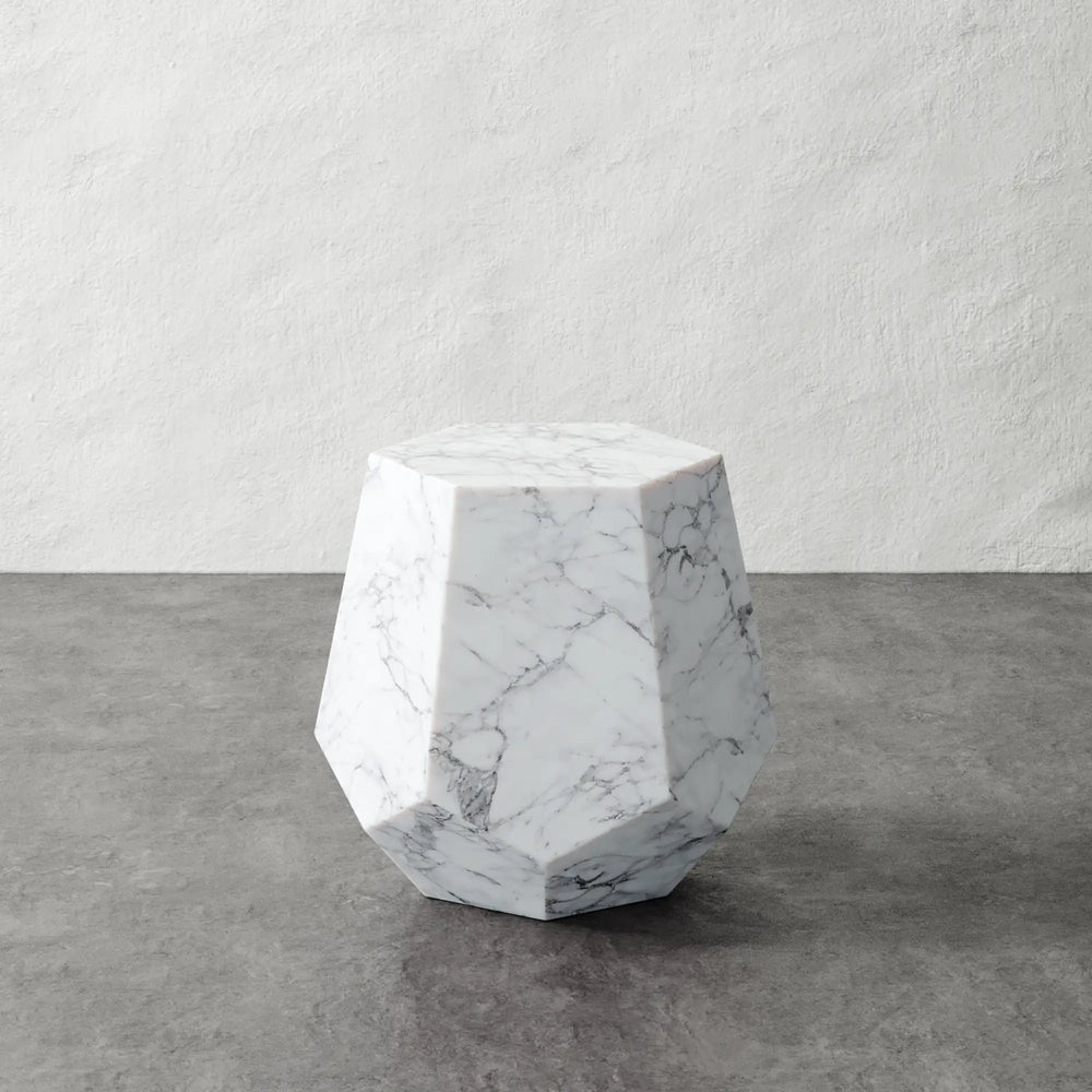 Lemieux et Cie Tessaire Faux Marble Accent Table in white resin with gray veining, geometric faceted side table on stone floor against textured plaster wall.