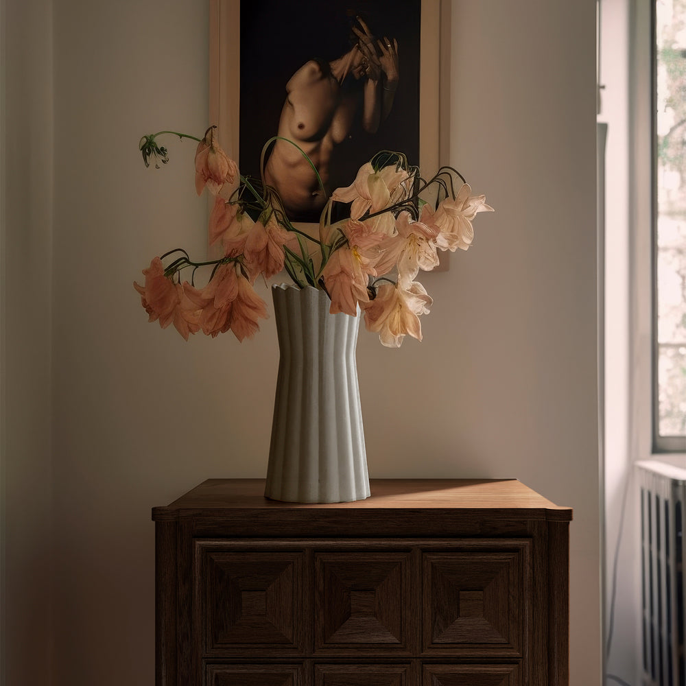 Lemieux et Cie Gaspard Vase with fluted form holding blush flowers, styled on dark wood cabinet below framed artwork in neutral-walled room with soft daylight.