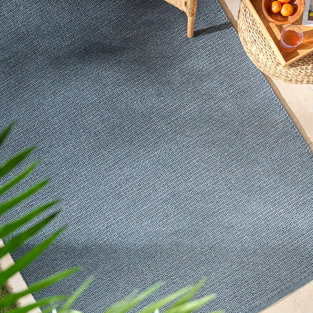 Lemieux et Cie Cordage Hand Woven Outdoor Rug in blue, diagonal weave pattern, shown on outdoor patio with woven chair and natural light.