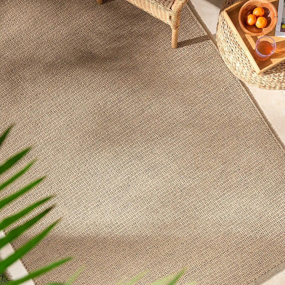 Lemieux et Cie Cordage Hand Woven Outdoor Rug in tan, flatweave rug on outdoor patio with woven furniture and warm light.