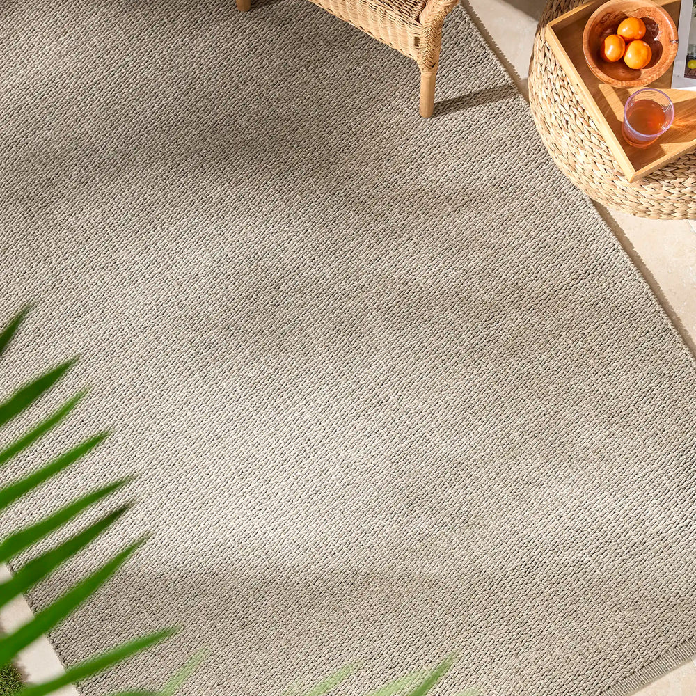 Lemieux et Cie Cordage Hand Woven Outdoor Rug in grey, textured diagonal weave rug shown with rattan chair and woven tray in warm sunlight.