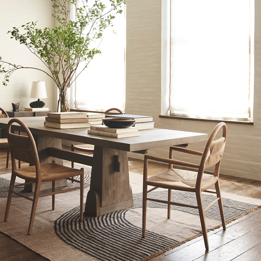Lemieux et Cie Clichy Dining Table in wire-brushed acacia with driftwood finish, dining table styled with woven chairs on striped rug in sunlit dining room with white walls and wood floors.