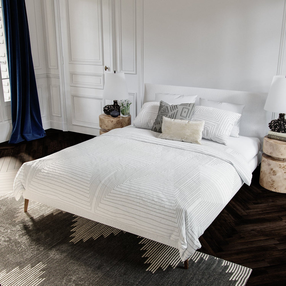 Lemieux et Cie Mondoro Quilt Collection in white cotton with geometric linear quilting, duvet and shams styled on upholstered bed in bedroom with white paneled walls and dark wood floors.