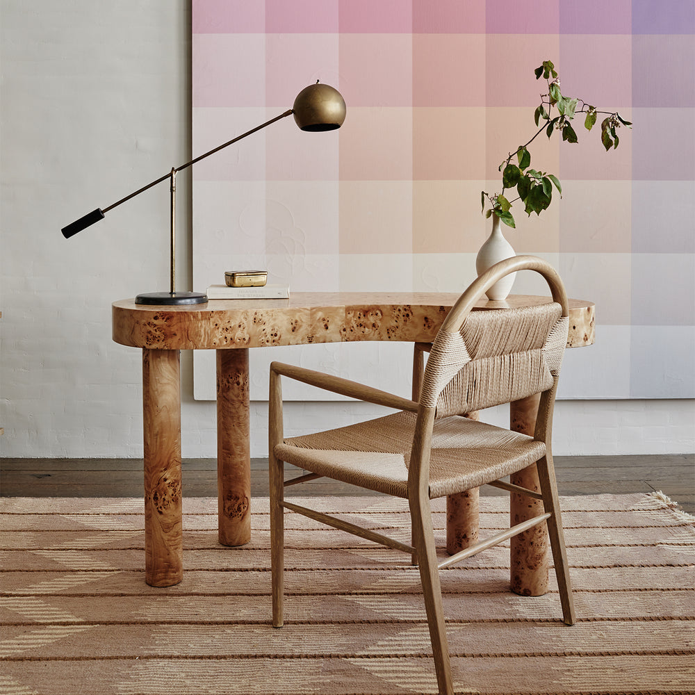 Lemieux et Cie Pernelle Dining Arm Chair in oak with woven rush seat, styled with burl wood table and brass task lamp against soft pink tiled wall.
