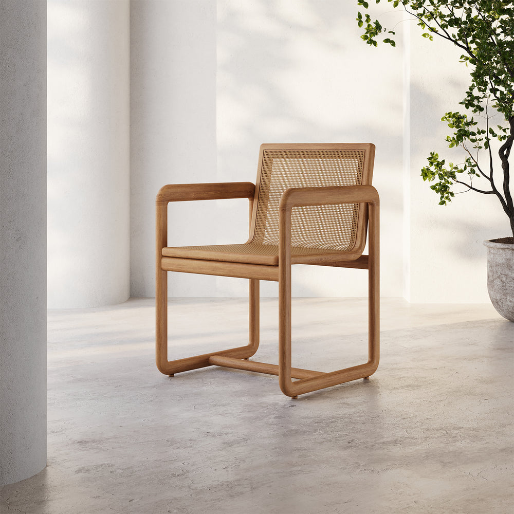 Lemieux et Cie Dock Teak Dining Armchair with woven cane panels in solid teak frame, shown on concrete floor in dappled sunlight.