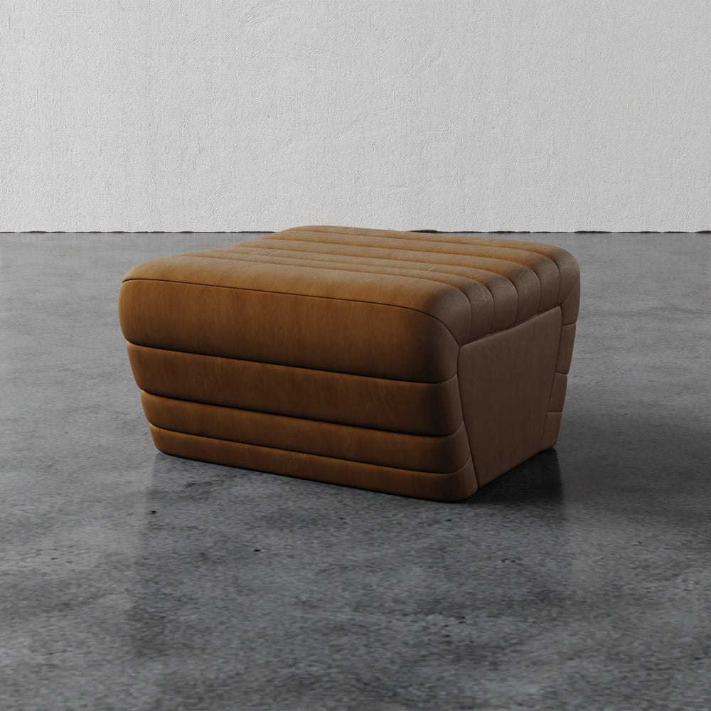 Lemieux et Cie Brunoy Leather Ottoman in warm tobacco, channeled leather ottoman with rounded sculptural form, shown on concrete floor.