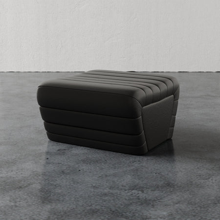 Lemieux et Cie Brunoy Leather Ottoman in charcoal with horizontal channel tufting, low-profile ottoman on stone floor against textured plaster wall.