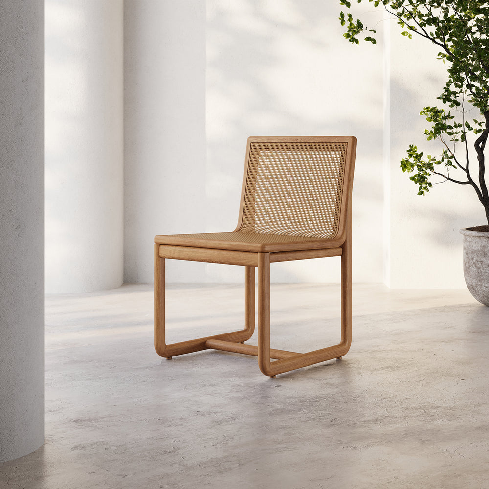 Lemieux et Cie Dock Dining Chair in solid teak with woven cane panel seat and back, outdoor dining chair on concrete patio with dappled sunlight.