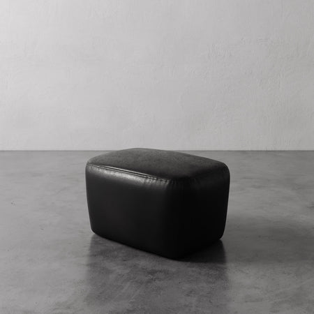 Lemieux et Cie Aurélie Leather Ottoman in black, rounded low-profile ottoman on polished concrete floor against textured plaster walls.