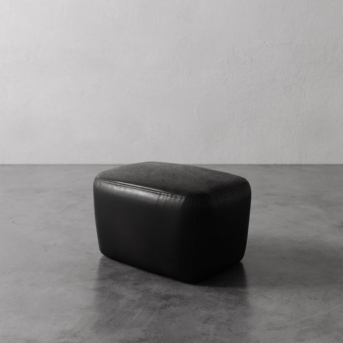 Lemieux et Cie Aurélie Leather Ottoman in black, rounded low-profile ottoman on polished concrete floor against textured plaster walls.