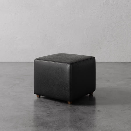 Lemieux et Cie Fabien Square Ottoman in black leather, low-profile cube form on dark legs, shown on concrete floor against textured plaster walls.