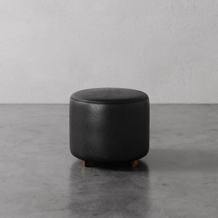 Lemieux et Cie Fabien Round Ottoman in black leather, cylindrical upholstered ottoman on warm wood legs, shown on polished concrete floor against textured plaster walls.