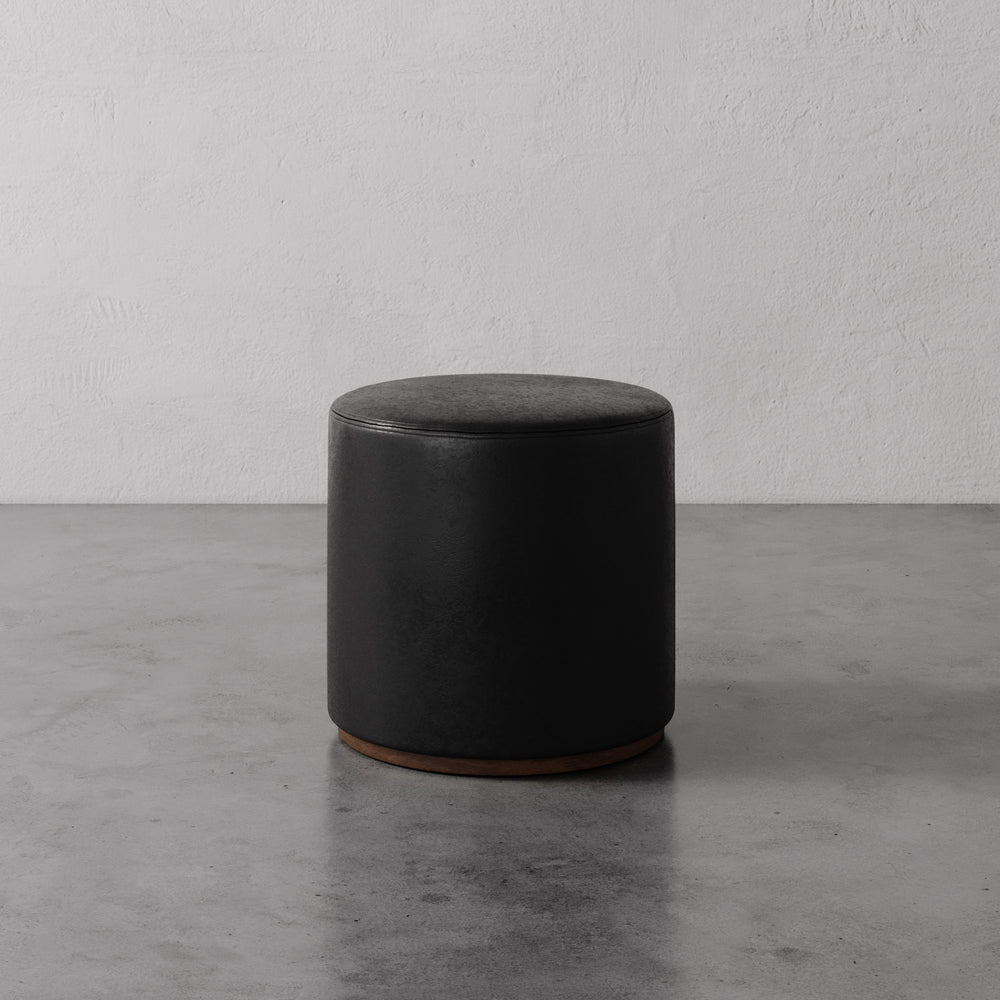 Lemieux et Cie Sophie Round Ottoman in black leather, cylindrical ottoman with warm wood base, shown on concrete floor against textured plaster wall.