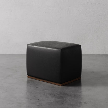 Lemieux et Cie Sophie Leather Rectangular Ottoman in dark leather with wood base, low-profile ottoman on concrete floor against textured plaster walls.