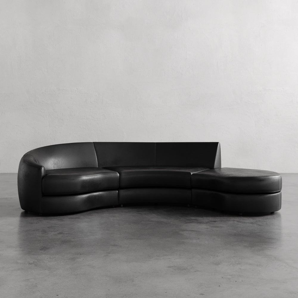 Lemieux et Cie Rulmann 3-Piece Left Sectional in black leather, curved sofa with rounded chaise, shown on concrete floor against white plaster wall.