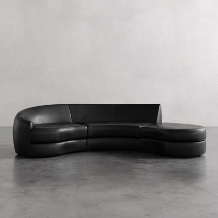 Lemieux et Cie Rulmann 3-Piece Left Sectional in black leather, curved sofa with rounded chaise, shown on concrete floor against white plaster wall.