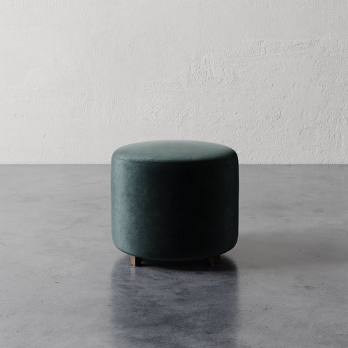 Lemieux et Cie Fabien Round Ottoman in deep green upholstery, low cylindrical form on turned legs, set against textured plaster wall and concrete floor.