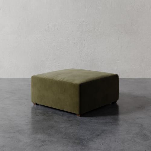 Lemieux et Cie Fabien 36" Square Ottoman in olive green upholstery, low-profile ottoman on dark stained legs, shown on concrete flooring against textured plaster walls.