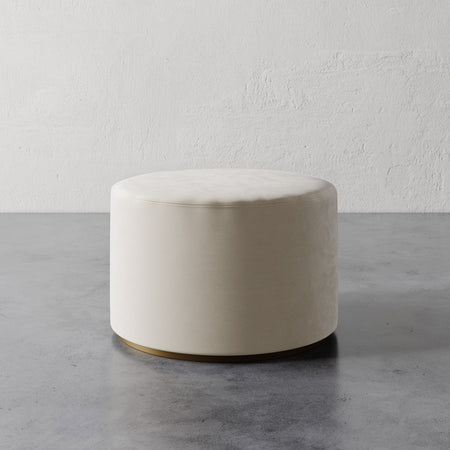 Lemieux et Cie Sophie Round Ottoman in ivory upholstery with brass base detail, cylindrical ottoman on concrete floor against textured plaster wall.